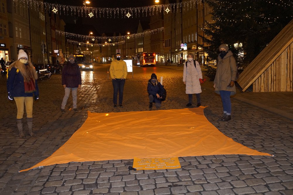 Mehrere Frauen stehen in der abends erleuchteten Landshuter Altstadt hinter einem großen orangen Tuch, das auf dem gepflasterten Boden ausgebreitet ist.