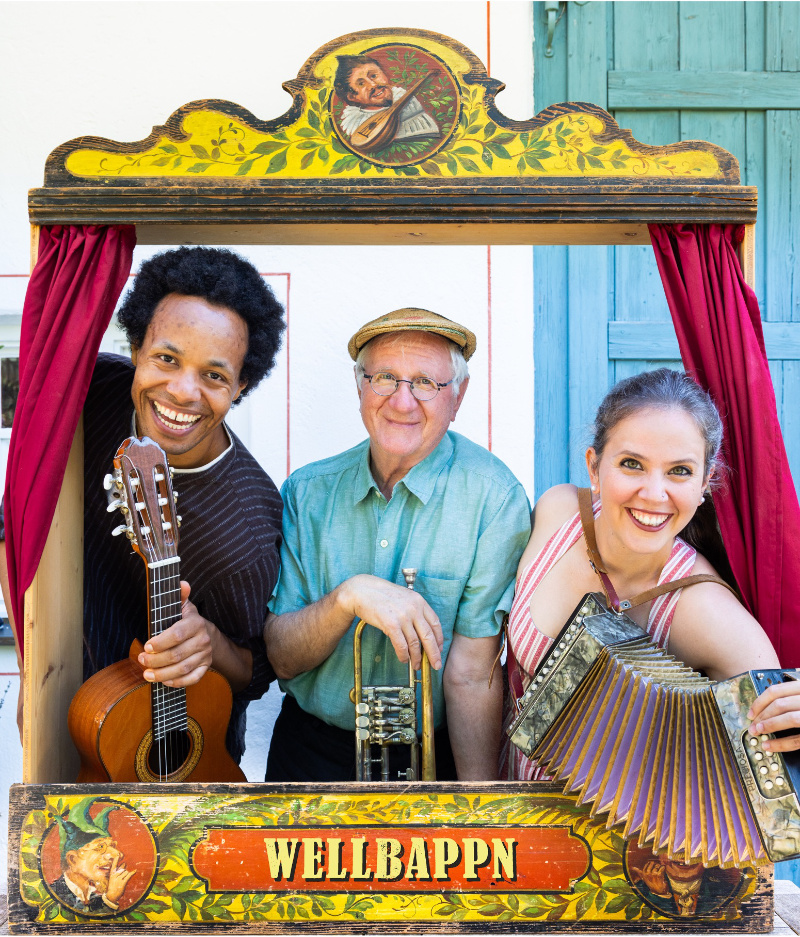 Hans Well, Sarah Well und Komale schauen mit Instrumeneten (Gitarre, Trompete, Ziehharmonika) aus einem alten Kaspertheater heraus