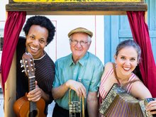 Hans Well, Sarah Well und Komale schauen mit Instrumeneten (Gitarre, Trompete, Ziehharmonika) aus einem alten Kaspertheater heraus