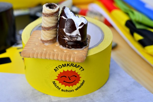 Auf einer gelben Kleberolle mit "Atomkraft? nicht schon wieder"-Logo liegt ein Butterkeks, auf dem eine Waffelrolle einen Kamin darstellt und ein angebissener Schaumzuckerkuß den Reaktorblock