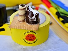 Auf einer gelben Kleberolle mit "Atomkraft? nicht schon wieder"-Logo liegt ein Butterkeks, auf dem eine Waffelrolle einen Kamin darstellt und ein angebissener Schaumzuckerkuß den Reaktorblock