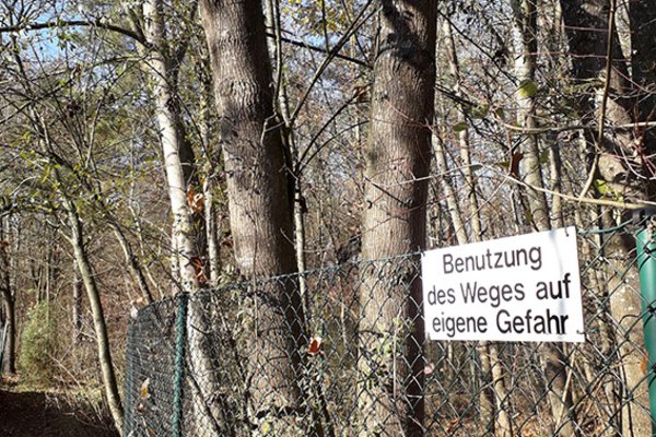 schmaler Fußweg durch mit Bäumen und Sträuchern bewachsene Au mit Zaun und Schild "Betreten des Weges auf eigene Gefahr"