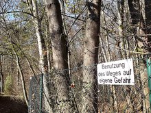 schmaler Fußweg durch mit Bäumen und Sträuchern bewachsene Au mit Zaun und Schild "Betreten des Weges auf eigene Gefahr"