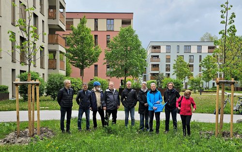 sehr viele Menschen stehen bei zwei frisch gepflanzten Bäumen. Willi Forster streicht auf der Gießkanne eine Zahl ab