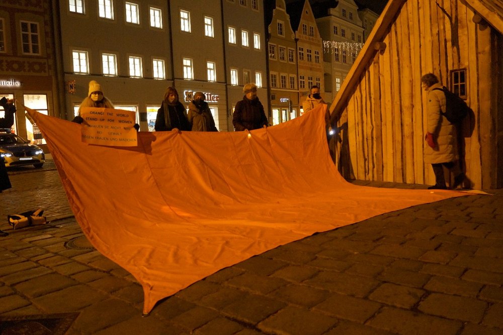 Mehrere Frauen stehen in der abends erleuchteten Landshuter Altstadt neben der Weihnachtskrippe hinter einem großen orangen Tuch, eine hält ein oranges Schild in der Hand mit der Aufschrift "Orange the world - stand up for women. Schauen Sie nicht weg bei Gewalt gegen Frauen und Mädchen". Im Hintergrund erleuchtete Fenster und ein Polizei-Auto.