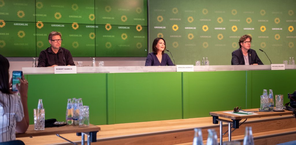 Robert Habeck, Annalena Baerbock und Michael Kellner sitzen vor einer "Gruene.de"-Plakatwand hinter einem langen Tisch an Mikrofonen. Im Vordergrund leere Konferenztische und ganz links eine Frau, die die Szene mit einem Mobiltelefon aufnimmt. (Foto: Nils Leon Brauer)