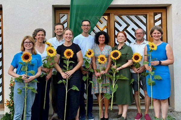 MdB Marlene Schönberger, Laura Pollard, Ferdinand Schoßer, Julia Oberland, Marek Cina, Dr. Simone Hartmann, MdL Mia Goller, Rudi Ruhland, Prof. Dr. Hannah Jörg mit Sonnenblumen in der Hand vor grüner Beach-Flag