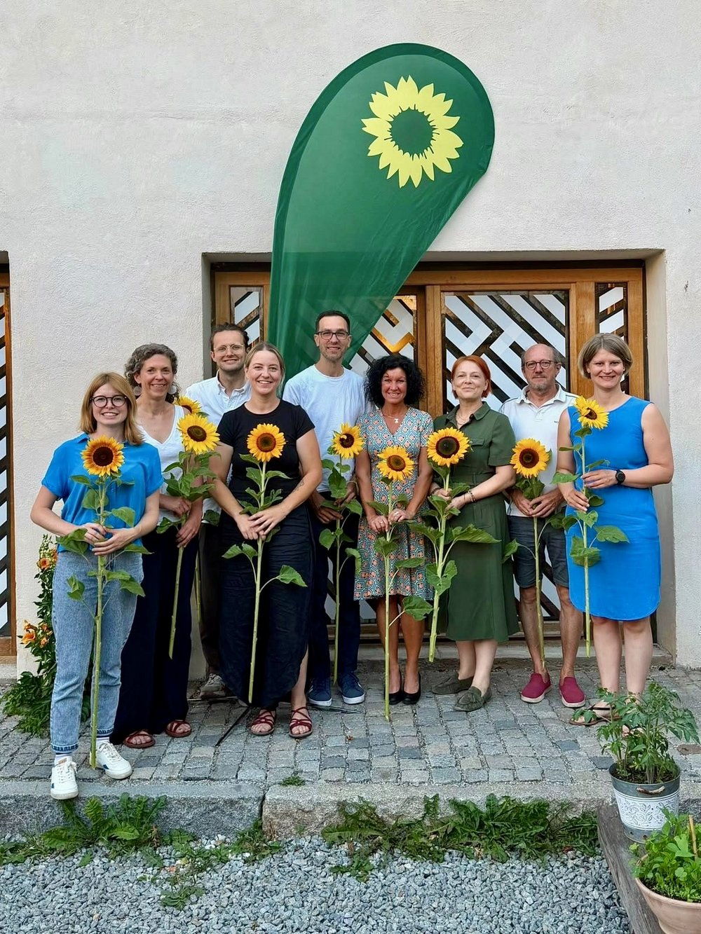 MdB Marlene Schönberger, Laura Pollard, Ferdinand Schoßer, Julia Oberland, Marek Cina, Dr. Simone Hartmann, MdL Mia Goller, Rudi Ruhland, Prof. Dr. Hannah Jörg mit Sonnenblumen in der Hand vor grüner Beach-Flag