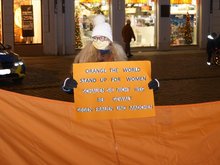 Eine Frau mit weißer Zipfelmütze und Mundschutz steht in der abends erleuchteten Landshuter Altstadt und hält ein oranges Schild in der Hand mit der Aufschrift "Orange the world - stand up for women. Schauen Sie nicht weg bei Gewalt gegen Frauen und Mädchen". Im Hintergrund erleuchtete Schaufensterscheiben und ein Polizei-Auto.