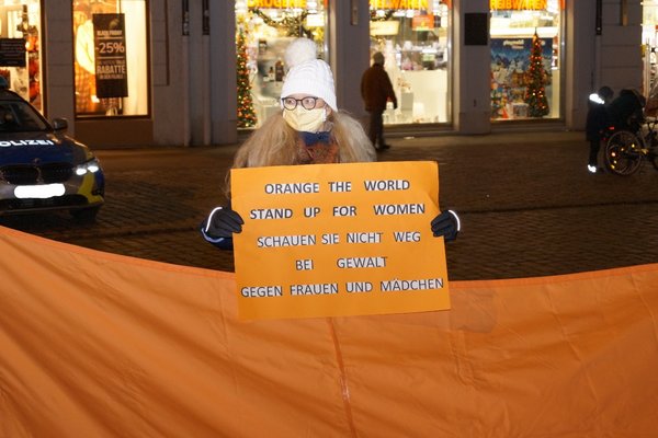 Eine Frau mit weißer Zipfelmütze und Mundschutz steht in der abends erleuchteten Landshuter Altstadt und hält ein oranges Schild in der Hand mit der Aufschrift "Orange the world - stand up for women. Schauen Sie nicht weg bei Gewalt gegen Frauen und Mädchen". Im Hintergrund erleuchtete Schaufensterscheiben und ein Polizei-Auto.