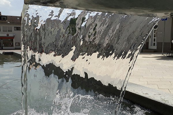 Foto eines von oben fließenden, breiten Wasserrstrahls aus einem Brunnen - von hinten fotografiert durch das Wasser hindurch