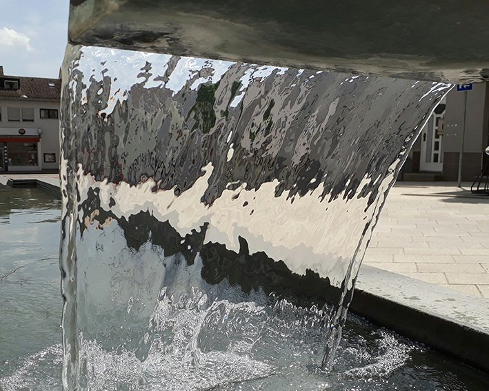 Foto eines von oben fließenden, breiten Wasserrstrahls aus einem Brunnen - von hinten fotografiert durch das Wasser hindurch