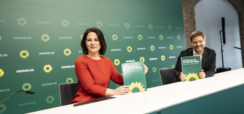 Annalena Baerbock und Robert Habeck sitzen an einem langen Tisch und halten den gedruckten Wahlprogramm-Entwurf in die Kamera. Dahinter eine "Gruene.de"-Wand (Foto: Dominik Butzmann)