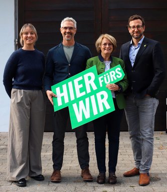 Das Spitzenquartett der grünen Stadtratsliste: Julia Oberland (Platz 3), Christoph Rabl (Platz 2), Iris Haas (Platz 1), Sebastian Bröckner-Kiss (Platz 4) mit einem grünen Schild mit der Aufschrift "HIER fürs WIR" vor einer hölzernen Eingangstüre