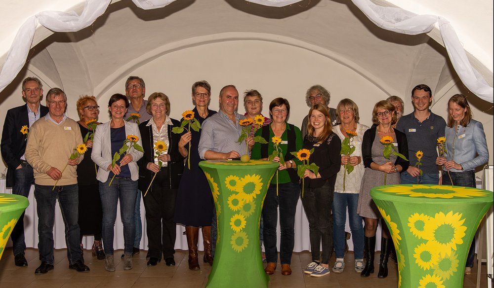 viele (17) freundliche Menschen mit Sonnenblumen in der Hand in einem Gewölbe unter weißen Stoffbahnen mit grünen Sonnenblumen-Stehtischen im Vordergrund: Grüne aus Stadt und Landkreis Landshut