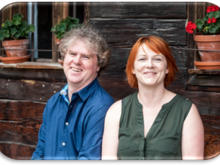 Markus Scheuermann und Mia Goller vor der Fassade eines alten hölzernen Bauernhauses mit blumengeschmückten Fenstern